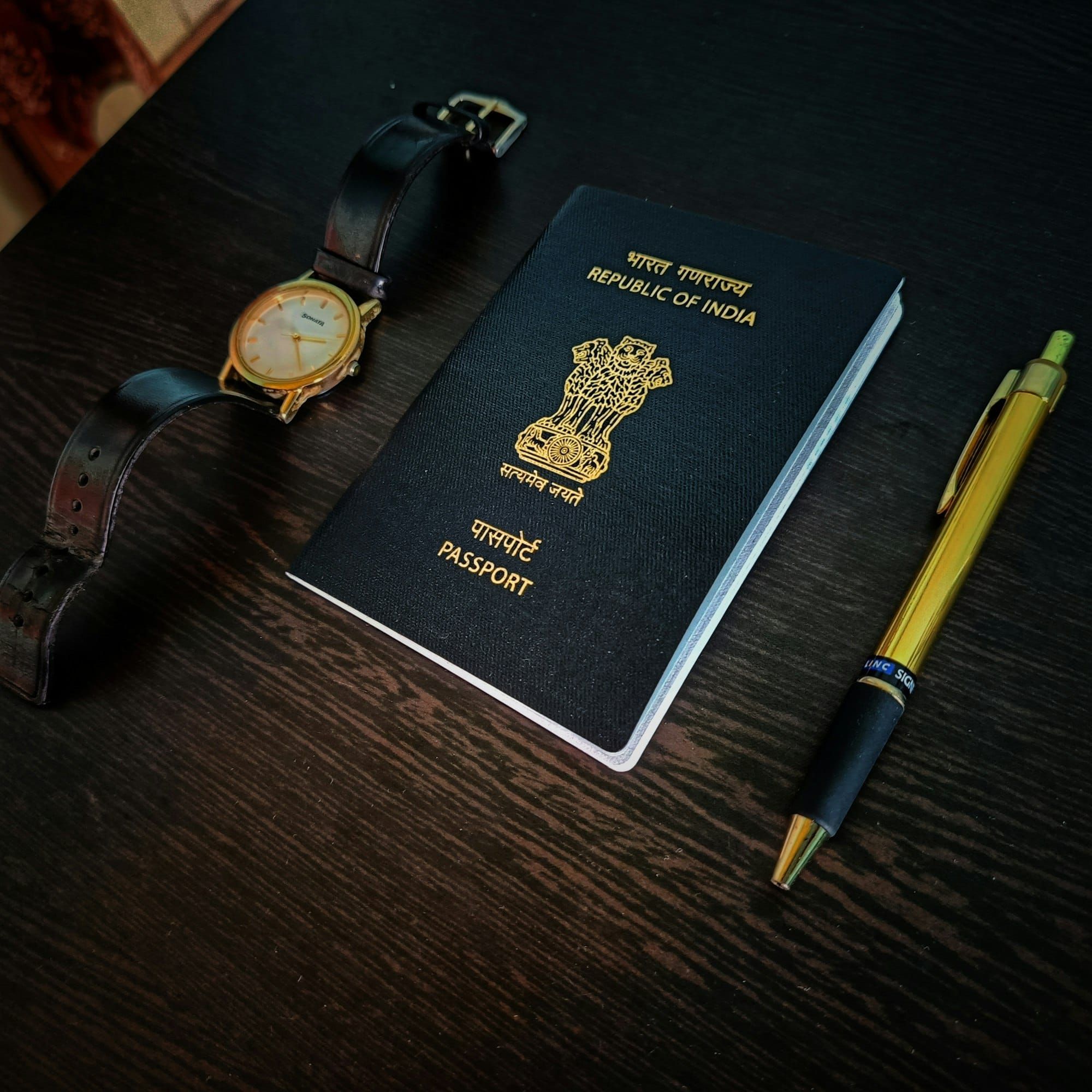 An Indian passport, a wristwatch and a pen on a dark wooden table