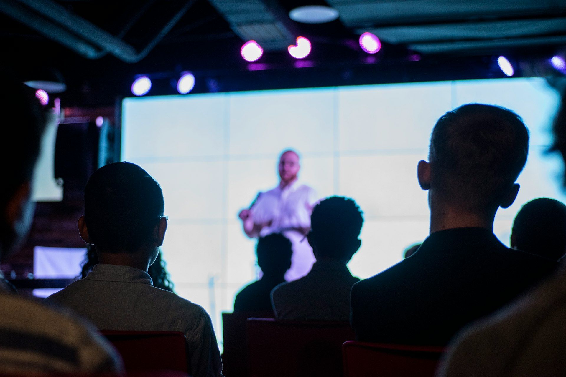 Audience members seen from behind watching a speaker in front of a bright stage screen
