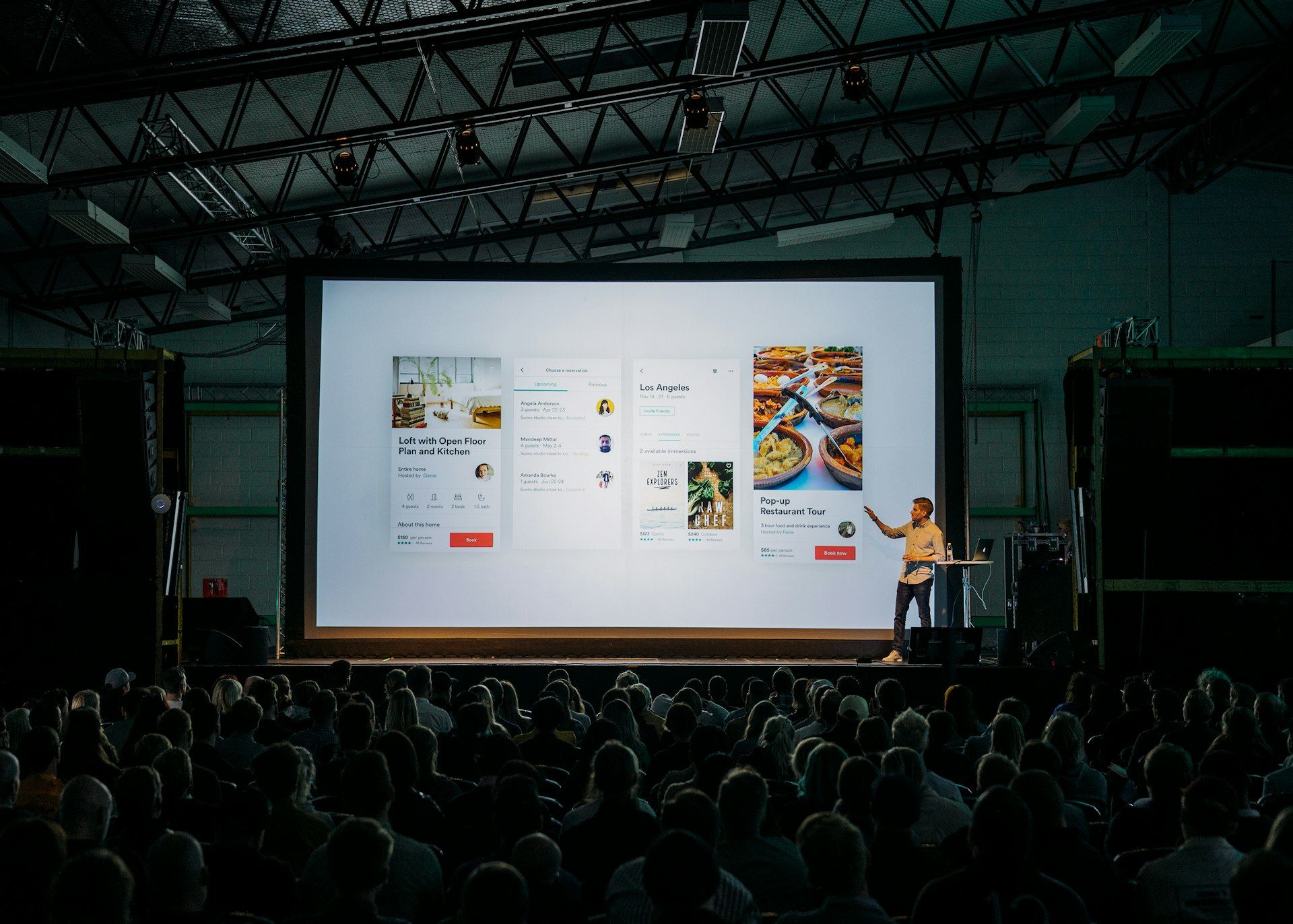 A presenter beside a large lit screen on a conference stage, the audience in silhouette